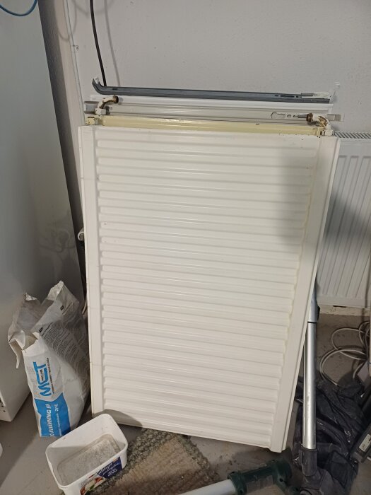 Gammalt element som lutar mot en vägg i en källare, bredvid en blåvit säck med byggmaterial och plaster.