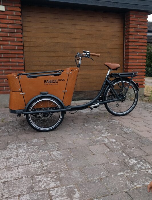 Lådcykel Babboe Curve i brunt och svart parkerad framför en garageport, sedd från sidan.