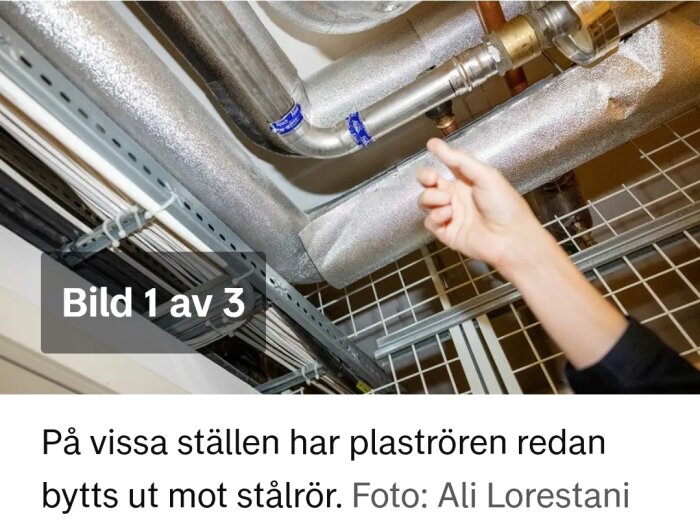 En hand pekar på stålklädda rör i ett innertak, omgivna av andra isolerade rör och kabeldragningar.