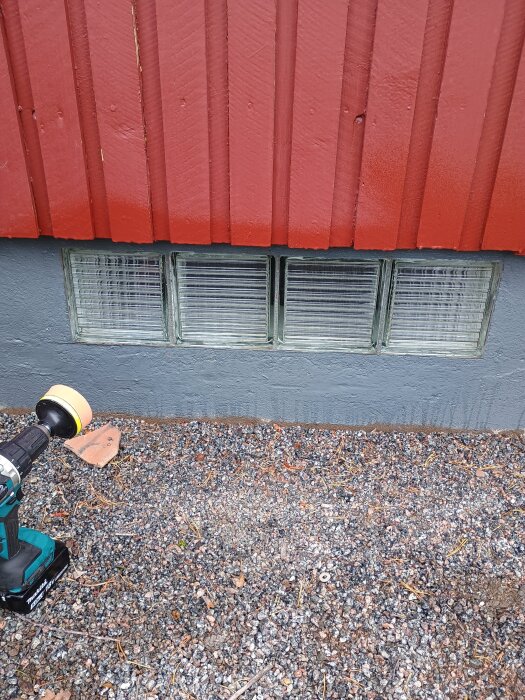 Fönster av glasblock vid källare, slipade och polerade, mot röd träfasad och grus, bredvid en elektrisk slipmaskin.