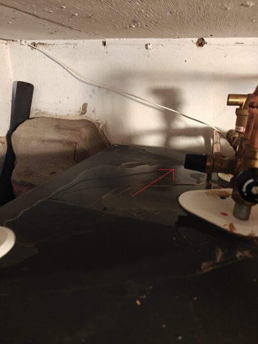 Vattenansamling ovanpå en pump med en pil som pekar mot en svart ventil i ett trångt utrymme under ett tak.