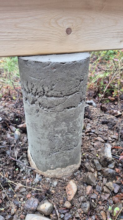 Betongplint med ojämn yta under en träbärlina, omgiven av jord och stenar. Möjliga kvalitetsproblem vid gjutning.