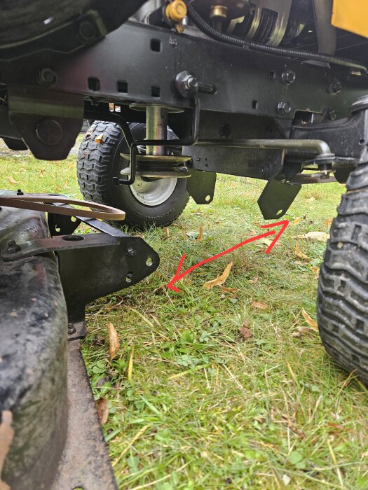 Underrede på gräsklippare med fokus på anslutning mellan klippaggregat och chassi, röda pilar markerar plats för saknat främre stag, gräsmatta i bakgrunden.