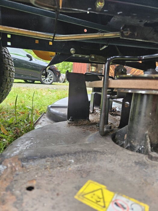 Vy under en traktorvisande ett gräsklippaggregat. Fokus ligger på metalldelar och remmar med gräs i bakgrunden.