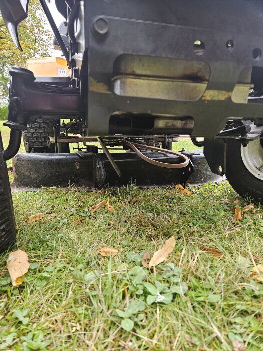 Traktor sedd underifrån med ny drivrem monterad. Klippaggregatet syns med ett saknat främre stag och ett framfäste med skador på en gräsmatta.
