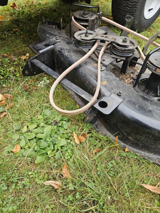 Närbild på en klippaggregatsmekanism med nyligen utbytt drivrem på gräsmattan, visar remmar och remskivor, brist på främre stag.