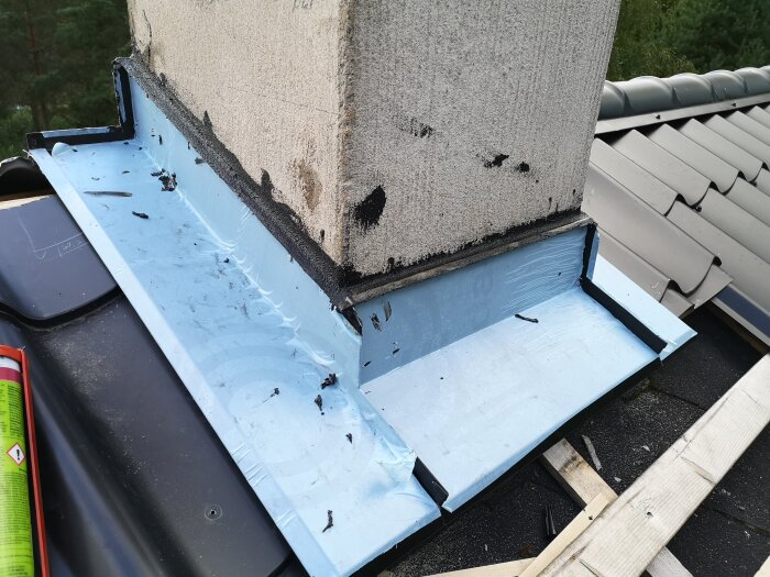 Plåtarbete runt en skorsten på ett svart tak med ljusblått skyddsmaterial och tätning i svart kitt.