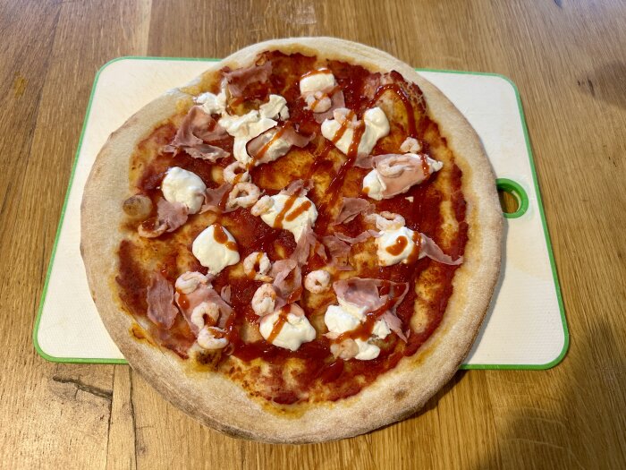 Hemgjord pizza med tomatsås, ost, skinka, scampi och röd sås på en skärbräda av trä.