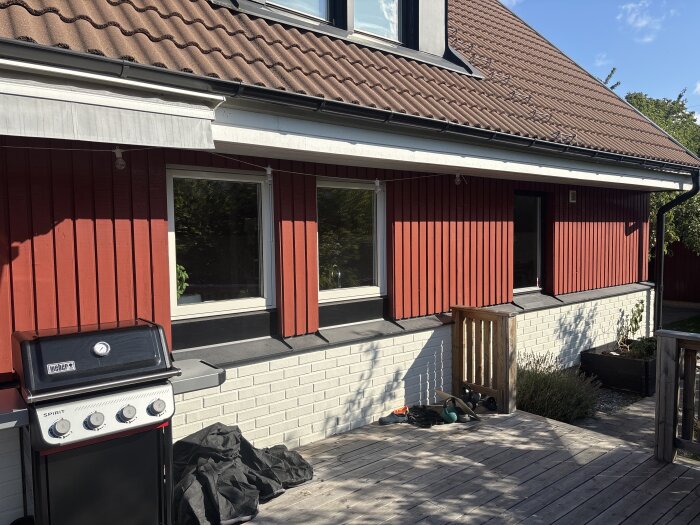 Rött träpanelsfasad med vitmålad tegelvägg, brun takpannor och grill på altanen.