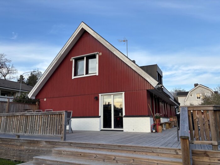 En röd villa med vitmålade tegelpartier och stora fönster, omgiven av ett trädäck, fotograferat under en klarblå himmel.