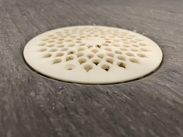 Vit, rund golvbrunnssil i plast med hål, monterad i duschgolv med mörkgrå matt yta.