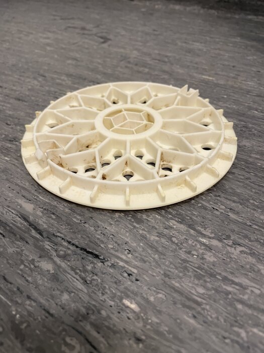 Vit plastgolvbrunnssil på ett gråaktigt golv med smuts i silens kanter.