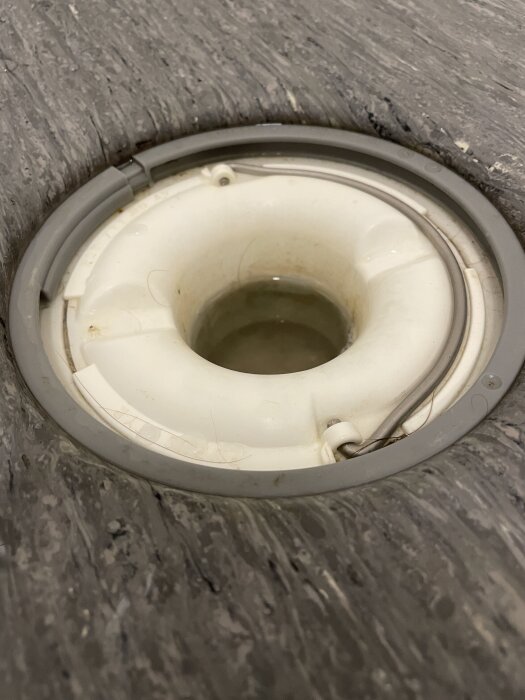 Bild av en golvbrunn i en dusch med hår och smuts runt kanten och på golvmattan. Problem med tätning och vattenlås synligt.