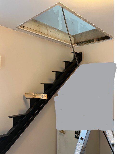 Metalltrappa monterad mot väggen med måttstock på trappstegen, öppning i taket ovanför.