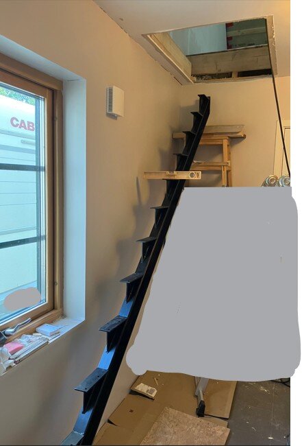 Svart balktrappa lutad mot vit vägg, leder till loftöppning. Ljusa väggar och fönster bredvid, arbetsområde med verktyg och material synligt.