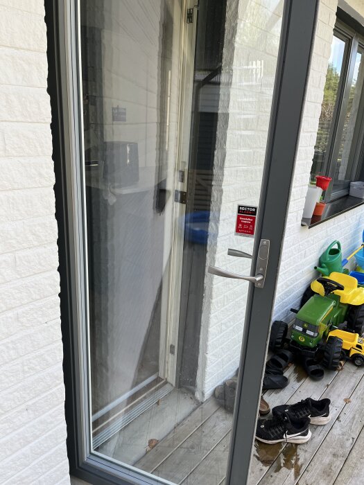 Grå dörrkarm i aluminium med glasfönster på uteplats, leksakstraktor, vattna kanna och skor syns i bakgrunden.