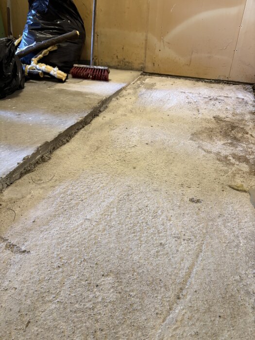 Betonggolv i garage med olika nivåer, delvis skrovlig yta, sopborste och svart sopsäck i bakgrunden.