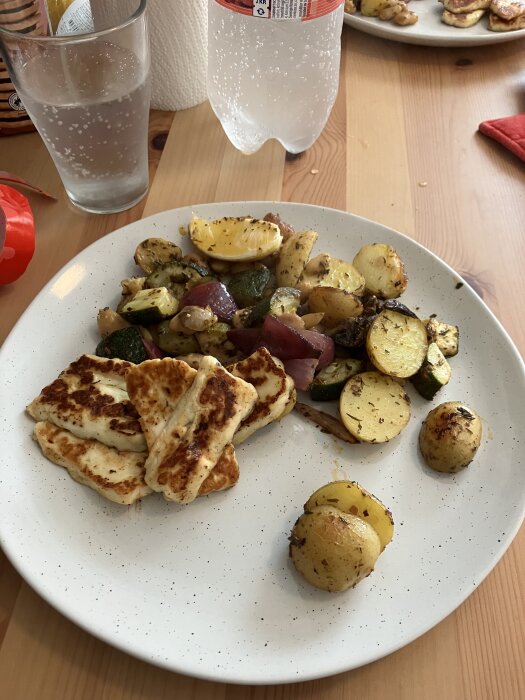 Tallrik med stekt halloumi, ugnsbakade potatisar, zucchini och svamp i grekisk marinad, samt ett glas och en flaska kolsyrat vatten bredvid.