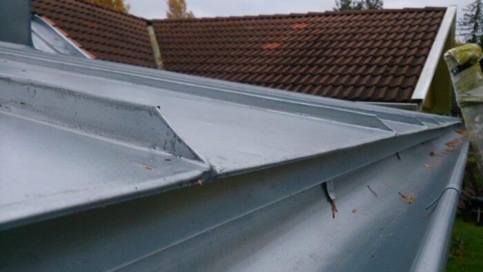 Dubbelfalsat aluminium-zink tak med rena vindskivor och avslut mot ränna, med tegelrött tak i bakgrunden.