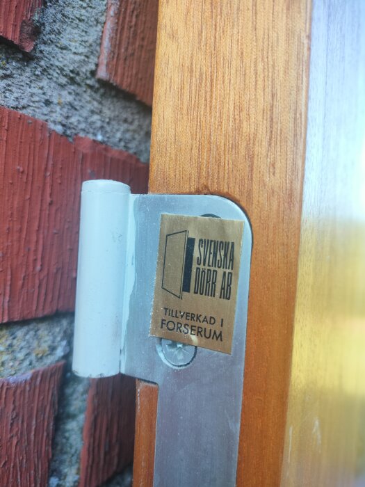 Närbild på gångjärn med etikett "Svenska Dörr AB, tillverkad i Forserum" monterat på en träyta intill en tegelvägg.