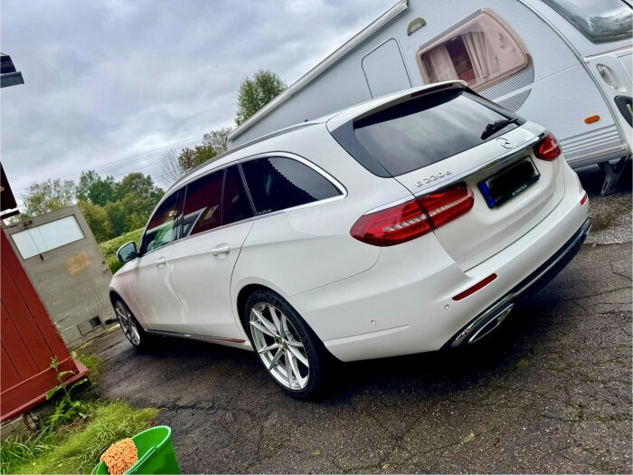 En vit Mercedes-Benz E220 stående framför en husvagn, parkerad på en asfalterad uppfart med grön hink i förgrunden.