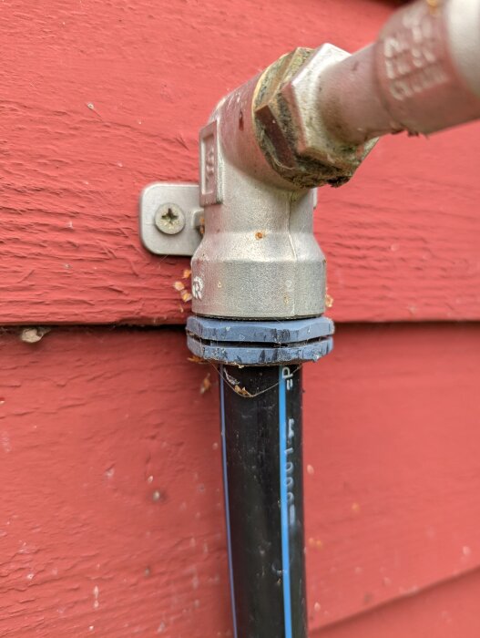 Vattenutkastare monterad på röd trävägg med ansluten svart vattenslang, förbunden med metallkoppling och skruvfäste.