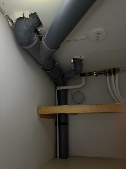 Bilden visar ett rum med synliga grå rör i taket och väggen, del av badrumsrenovering. Målningen runt rören är skadad, och träbräda syns.
