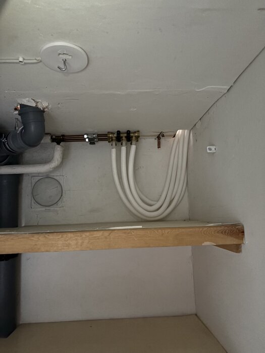 Rördragningar i badrum med vit vägg och en trälist synlig. Flera vita och gråa rör är kopplade till mässingsfästen på väggen.
