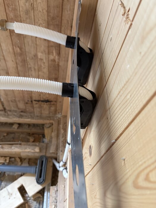 Rördragning i badrum med synliga vit korrugerade rör och träpanel i bakgrunden under pågående renovering.