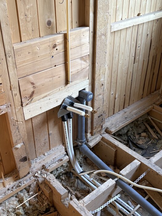 Rördragningar i trävägg under badrumsrenovering, med flera rör och ledningar synliga, och isoleringsmaterial på golvet.