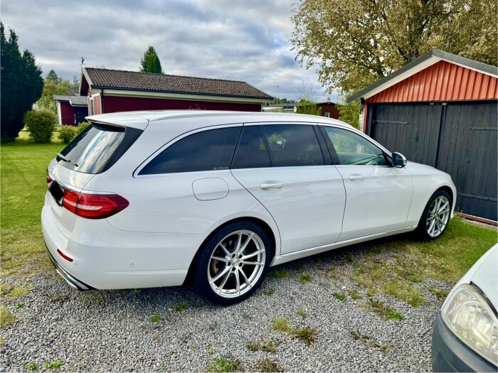 Vit Mercedes E220 parkerad på grus vid garage, omgiven av gräs och träd.