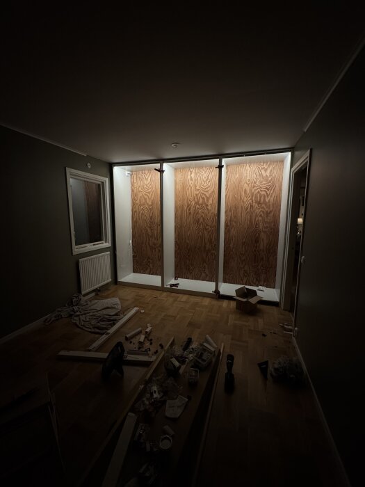 Rum under renovering med tre garderober med plywoodbaksida, oisolerad strömbrytare, verktyg och byggmaterial utspridda på golvet.