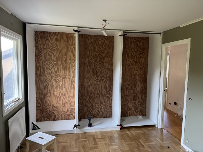 Tre PAX-garderober med plywood-baksidor under installation i ett rum med trägolv och gröna väggar, sett från ett fönster.