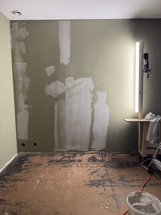 Vägg under renovering med spacklade ytor och en arbetslampa på stativ. Golvet är avskalat med synliga märken. En burk med spackel står på golvet.