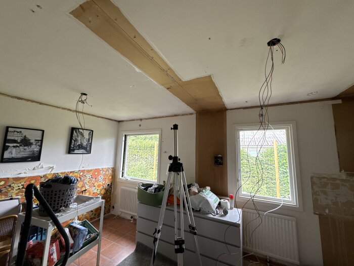 Rum under renovering med synliga elkablar i taket, verktyg, möbler och tapetserade väggar. Ett fönster och en dörr syns, samt målade väggar.