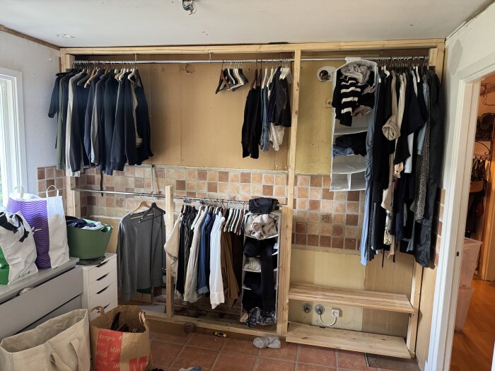 Oanvänd garderobsstruktur under byggnation med kläder hängande på klädstänger, korgar och klädhängare, samt tegelvägg i bakgrunden.