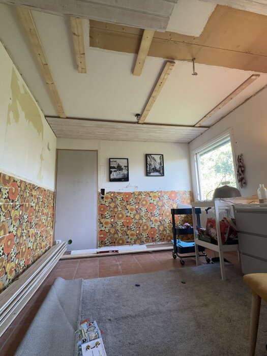 Rum under renovering med gammalt blommigt tapetmönster, synliga takbjälkar och arbetsmaterial spritt över golvet.