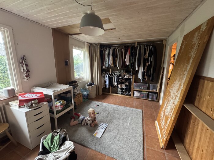 Barn leker på golvet i rum med öppen garderob fylld med kläder, lådor och en oinfäst dörrskiva lutad mot väggen; renoveringsdetaljer och leksaker synliga.