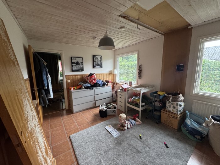 Rum i renovering med trätak, vitmålade väggar och bröstpanel. Inredning är dysfunktionell med oorganiserade föremål och barn som leker på golvet.