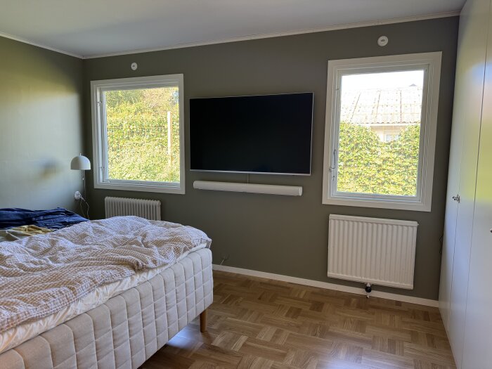Sovrum med två fönster, en väggmonterad TV och en soundbar, parkettgolv, beige säng och vita element.