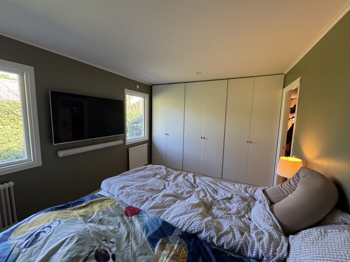Renoverat sovrum med dubbelsäng, vit garderob, och väggmonterad TV. Grönmålade väggar och två fönster visar dagsljus. Beige sängkudde och lampa på sidan.