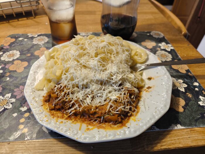 Tallrik med köttsås och tagliatelle toppat med riven Pecorino, serverad med ett glas dryck och en karaff på en blommig bordstablett.