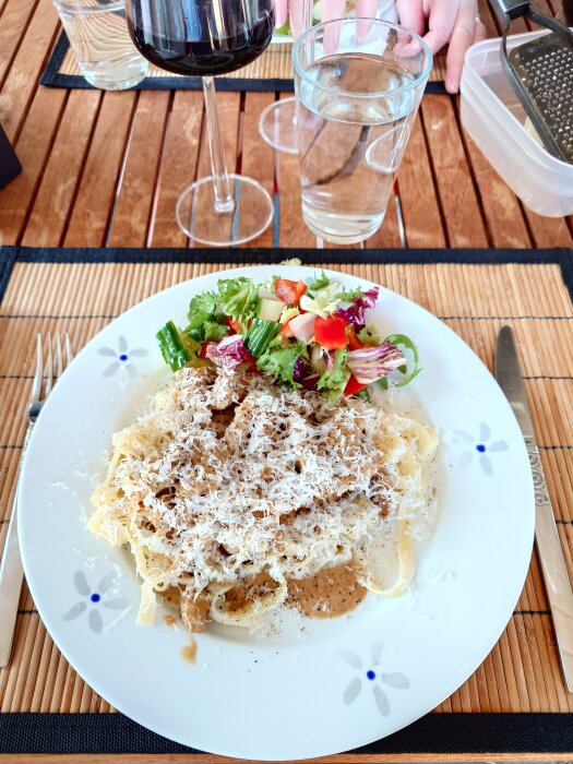 Tallrik med fläskfilépasta toppad med parmesan, svartpeppar och sallad. Vin och vatten i glas på bordet.