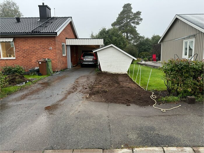 Uppfart med grävd mark, bil parkerad under carport, och linjer markerade för framtida avgränsning, intill två hus.