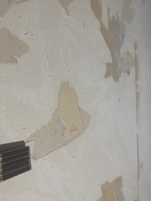 Vägg med delvis borttagen tapet, ojämn yta med flera lager synliga. Verktyg för tapetborttagning i nedre vänstra hörnet.