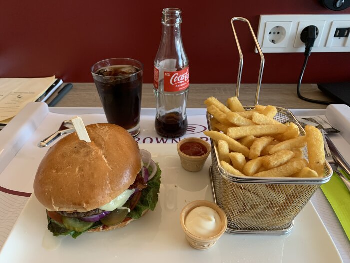 Hamburgare med sallad och lök, pommes frites i metallkorg, och cola i flaska och glas, serverat med ketchup och majonnäs på bricka.