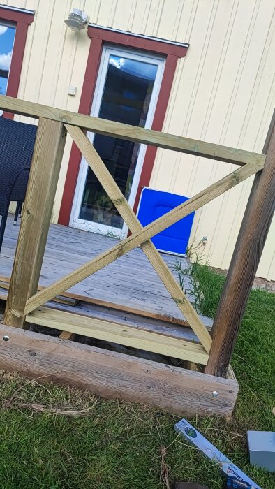 Trästaket under uppbyggnad med kryssmönster, delvis färdigt på en träaltan framför en gulmålad husvägg med vita fönster.