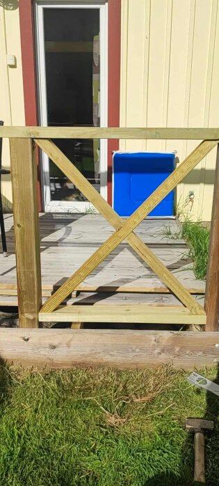 Trästaket med kryssmönster vid en altan framför en gul fasad och blå föremål, gräs och verktyg syns i förgrunden.