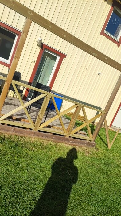 Altan med träkryssräcke under konstruktion, del av bygge runt husfasad. Solbelyst gräsmatta i förgrunden.