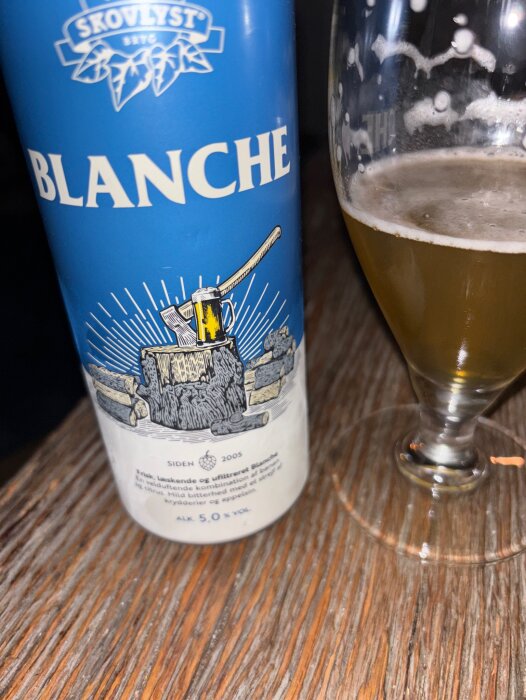 Blå burk med Skovlyst Blanche veteöl och ett glas öl på träbord.
